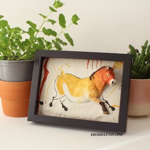 Puede incluir: Una obra de arte enmarcada con una ilustración de un caballo en tonos amarillos, naranjas y marrones, con contornos negros. La obra está en un marco negro y el fondo es de color beige claro. También se ven plantas en macetas.