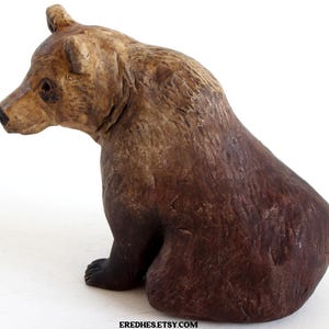 May include: A brown bear figurine with a sitting posture. The bear has a textured surface with varying shades of brown and tan. The bear's paws are black, and the figurine is set against a white background.