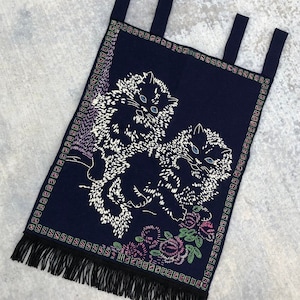 May include: A navy blue tapestry with a fringe border features two white embroidered cats with pink and green flowers. The tapestry has three loops at the top for hanging.