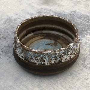 Pode incluir: Uma tigela de cerâmica artesanal com uma borda texturizada e salpicada. A tigela apresenta uma base marrom escura, um interior azul claro e uma borda texturizada branca e marrom. A tigela é redonda e tem uma aparência rústica e artesanal.