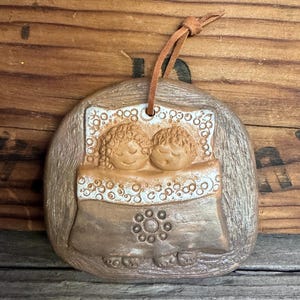 May include: A ceramic ornament featuring two figures in bed, with brown and beige tones. The figures are under a patterned blanket, with a leather cord for hanging. The ornament has a rustic, handmade appearance.