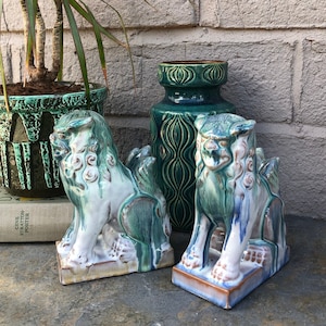 May include: Two green and white ceramic foo dog figurines with intricate details. The figurines are sitting on a brown surface in front of a brick wall.