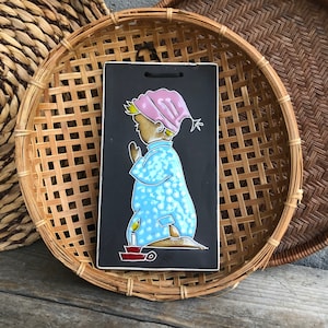 May include: A ceramic tile with a black background and a blue and white illustration of a child in a nightgown holding a candle. The child is wearing a pink hat and has a white halo.