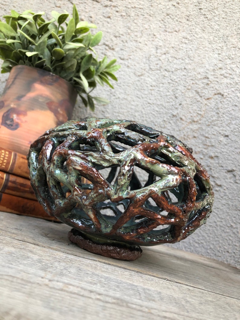 May include: A ceramic sphere with a lattice-like design in shades of brown, green, and white. The sphere is sitting on a wooden surface.