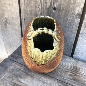 May include: A brown and yellow ceramic vase with a unique, irregular opening. The vase has a textured surface and a rustic appearance.
