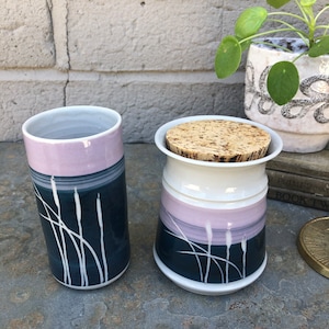 Puede incluir: Dos jarras de cerámica con un diseño azul oscuro y rosa claro. Las jarras tienen un diseño blanco de hierba alta con puntas blancas. La jarra de la derecha tiene una tapa de corcho.