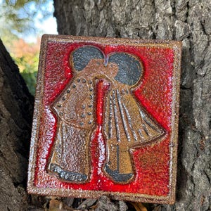 May include: Square ceramic tile with a red background and a brown border. The tile features a relief image of two figures kissing, with brown clothing and black hair. The figures are set against a textured red background.