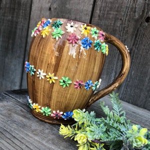 Puede incluir: Una jarra de cerámica marrón con asa, decorada con flores de colores. La jarra tiene un aspecto rústico y vintage y es perfecta para añadir un toque de encanto a cualquier cocina o comedor.