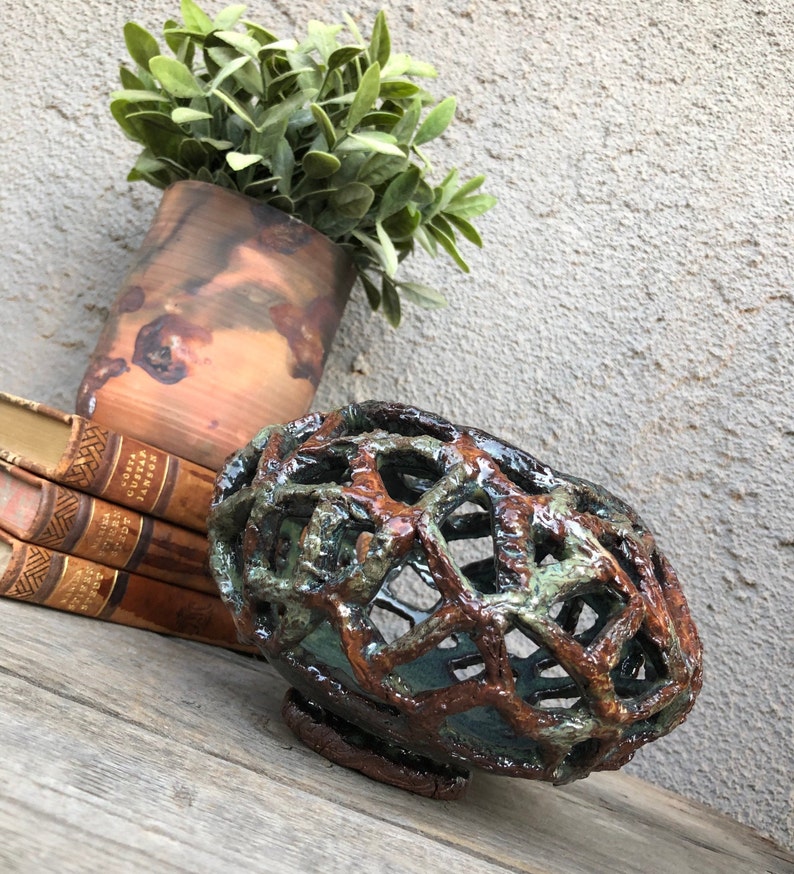 May include: A ceramic bowl with a unique, open-weave design. The bowl is glazed in shades of brown, green, and blue, giving it a rustic and earthy look.