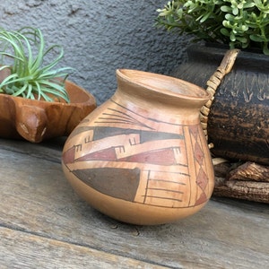 Puede incluir: Una pequeña maceta de cerámica pintada a mano con un diseño geométrico en tonos marrones, rojos y blancos. La maceta está sobre una superficie de madera.