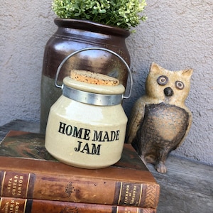 May include: A ceramic jar with a metal lid and a cork stopper. The jar is labeled "Home Made Jam". The jar is sitting on top of a stack of three leather-bound books. A ceramic owl figurine is to the right of the jar.