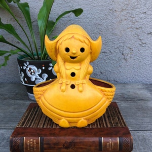 Könnte beinhalten: Ein gelber Keramik-Pflanztopf in Form eines Mädchens mit einem Häubchen. Der Topf hat zwei Löcher an der Seite für die Drainage.