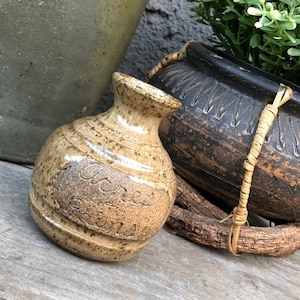 Puede incluir: Un pequeño jarrón de cerámica marrón y beige con cuerpo redondeado y cuello estrecho. El jarrón tiene una inscripción manuscrita que dice "Jackie's Pottery".