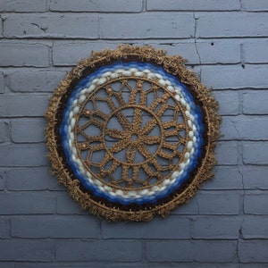 May include: A brown and blue woven wall hanging with a circular design. The center of the design is a large, intricate starburst pattern. The outer ring of the design is a series of concentric circles in shades of blue and white.