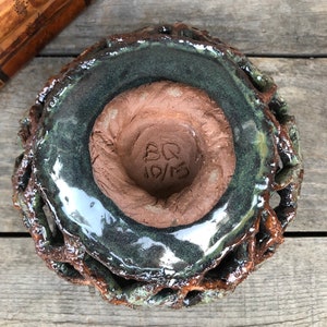 May include: A green glazed ceramic bowl with a brown clay base. The bowl has a unique, textured design and a signature that reads "BQ 10/13".