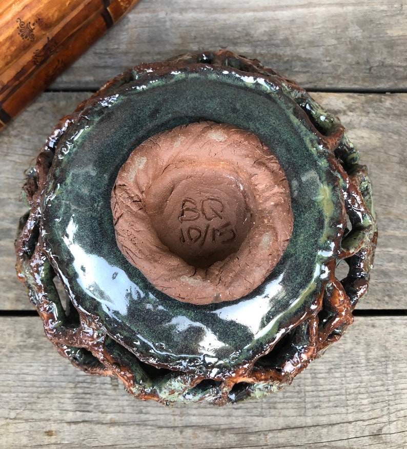 May include: A green glazed ceramic bowl with a brown clay base. The bowl has a unique, textured design and a signature that reads "BQ 10/13".