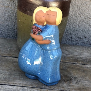 Puede incluir: Una figurita de cerámica de dos personas abrazándose, ambas con cabello rubio y vestidas con vestidos azules. La mujer de la izquierda sostiene un ramo de flores rosas.