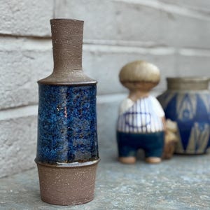 May include: A ceramic vase with a dark brown top and base, and a textured, cylindrical blue body. The vase is displayed with two other ceramic figurines, one of a person and another vase with a blue and white pattern.