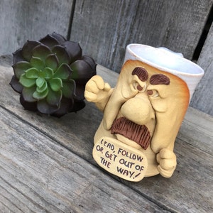 以下が含まれることがあります： 不機嫌な表情をした漫画の顔の形をしたセラミックマグカップ。マグカップには茶色の口ひげと眉毛、そして「Lead, Follow or Get Out of the Way!」と書かれた看板があります。