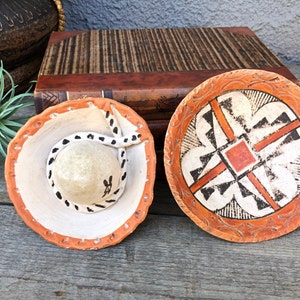 Pode incluir: Dois pequenos pratos de cerâmica com designs inspirados nos nativos americanos. Um prato é redondo com um padrão geométrico em preto, branco e laranja. O outro prato tem a forma de um sombrero com um design em preto e branco.