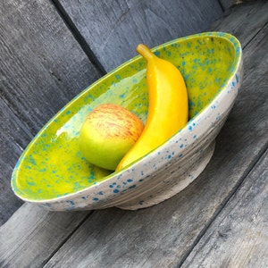 May include: A green and white ceramic bowl with blue speckles, filled with a yellow banana and a green apple.