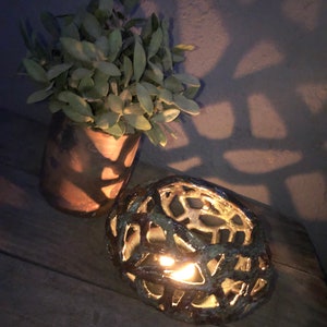 May include: A ceramic candle holder with a geometric design. The holder is brown and green with a light inside. The candle holder is sitting on a wooden surface.