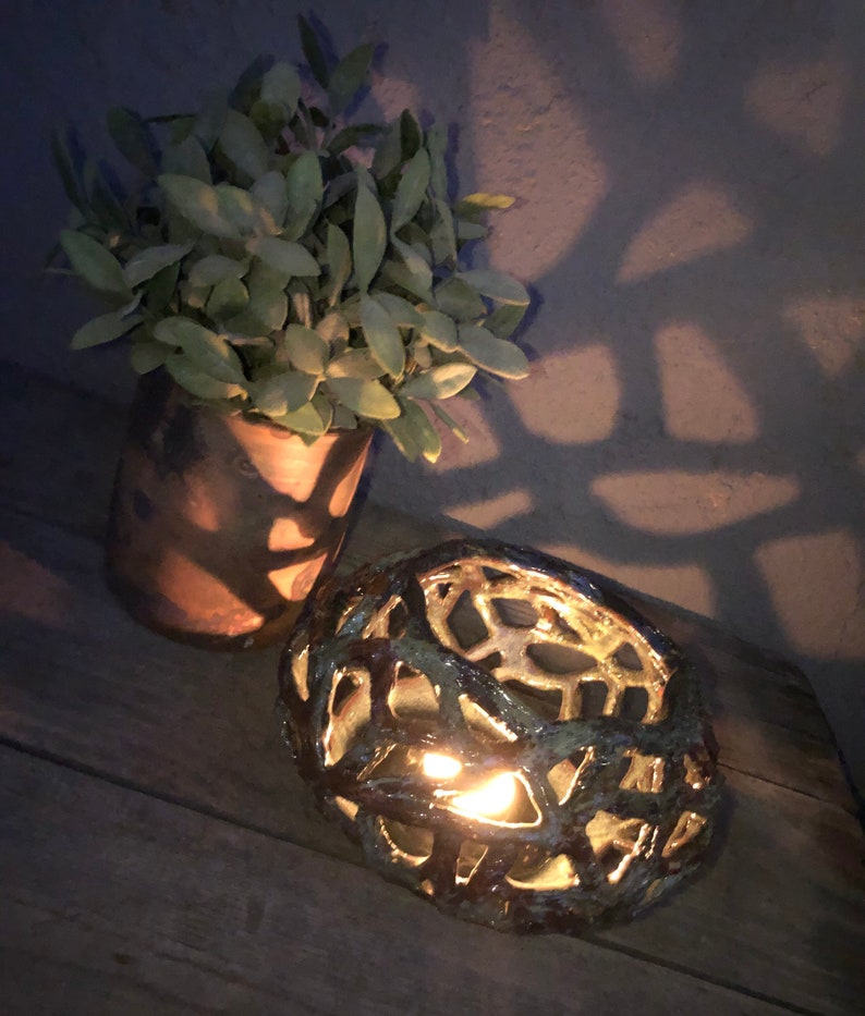 May include: A ceramic candle holder with a geometric design. The holder is brown and green with a light inside. The candle holder is sitting on a wooden surface.