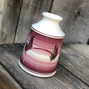 May include: A small, white ceramic vase with a pink and white floral design. The vase has a rounded base and a narrow neck.