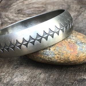 May include: A silver bangle bracelet with a geometric pattern etched into the surface. The bracelet is resting on a rough, brown rock.