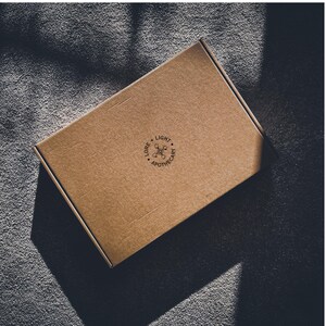 May include: A rectangular, brown cardboard box with the text "LORE + LIGHT APOTHECARY" printed in black. The box is positioned at an angle, with shadows cast across the surface. The box is on a gray, textured surface.