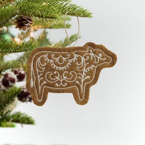 May include: A brown felt cow ornament with white embroidered detailing, hanging from a Christmas tree. The ornament features an intricate floral design within the cow's silhouette, adding a festive touch to holiday decor.