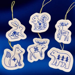 May include: Set of six white fabric ornaments with blue embroidered animal designs: a rabbit, squirrel, deer, fox, moose, and bear. Each ornament has a yellow string for hanging. The ornaments are on a blue background.