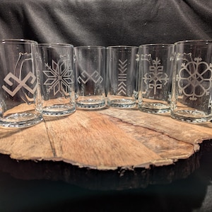 Puede incluir: Seis tazas de vidrio transparente con asas, cada una con un diseño grabado diferente. Las tazas están dispuestas en una fila sobre una superficie de madera.