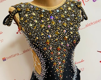 May include: A black leotard with a gold and clear rhinestone design. The leotard has a scoop neck and a low back. The leotard is decorated with a variety of rhinestones, including gold, clear, and iridescent.