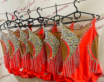 May include: A group of six leotards with a red base, brown and white mesh, and rhinestones. The leotards have a fringe trim and are hanging on black hangers.