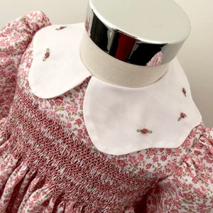 May include: A white smocked dress with a pink floral print and a white Peter Pan collar with embroidered green leaves and pink flowers.
