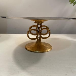 May include: A gold metal cake stand with a round glass top and a decorative base. The base is made of intertwined circles that resemble bamboo.