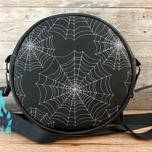 May include: Black round crossbody bag featuring a white spiderweb design. The bag has a black adjustable strap and silver-tone hardware. The bag is made of a durable material and is perfect for carrying essentials.