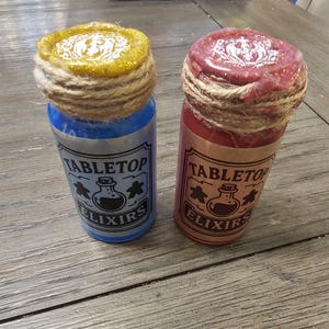 May include: Two glass bottles with corks wrapped in twine. One bottle is blue with a gold top, the other is red with a red top. Both bottles have a label that reads "Tabletop Elixirs" with a potion bottle and star icons.