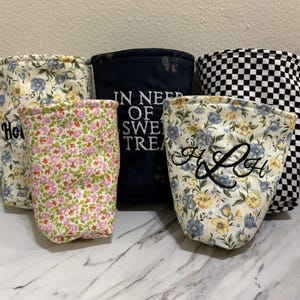 Könnte beinhalten: Fünf Stoffschalen-Wärmer mit verschiedenen Mustern. Einer hat ein blau-gelbes Blumenmuster mit Text, einer ein kleines rosa-grünes Blumenmuster, einer ist dunkelblau mit dem Text IN NEED OF SWEET TREATS, einer hat ein blau-gelbes Blumenmuster mit gestickten Initialen HLH und einer ein schwarz-weißes Schachbrettmuster.