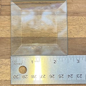May include: A clear, square, glass display box with a ruler showing inches. The box is approximately 2 inches wide. The box is empty and sits on a wooden surface. The ruler is silver with black markings.