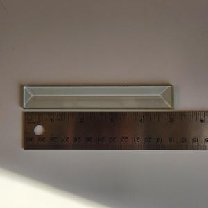 May include: A rectangular, light-colored glass tile, approximately 5 inches long, is positioned above a metal ruler. The ruler displays inch measurements. The tile has beveled edges and a reflective surface.