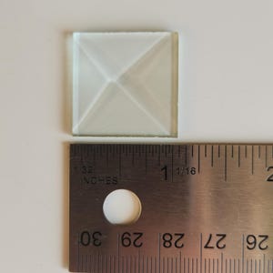May include: A clear, square glass tile with a pyramid design, measuring approximately 1 inch. A metal ruler with inch markings is placed next to the tile on a white surface.
