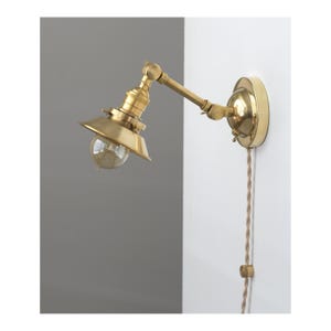 May include: A brass wall-mounted lamp with an adjustable arm and a conical shade. The lamp features a clear bulb and a braided cord. The fixture is mounted on a white wall with a gray background.