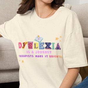 Puede incluir: Una camiseta de manga corta color crema con el texto DYSLEXIA IS A JOURNEY THERAPISTS MAKE IT BRIGHTER en letras coloridas y estampadas. Un pequeño icono de un libro abierto y estrellas amarillas están impresos sobre y alrededor del texto.