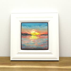 May include: A framed oil painting of a sunset over water. The sky blends from blue to orange and yellow, mirrored in the water. The white frame surrounds the artwork, displayed on a wooden surface.