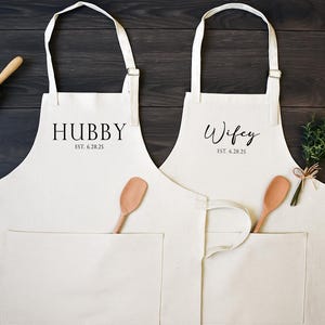 May include: Two off-white kitchen aprons with adjustable neck straps and front pockets. One apron features the black text HUBBY EST. 6.28.25, and the other features the black text Wifey EST. 6.28.25. Each apron includes a wooden spoon tucked into the pocket.