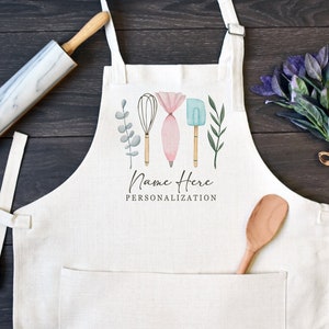May include: A cream-colored apron featuring a central graphic of a whisk, a piping bag, a spatula, and green foliage. The text "Name Here" and "PERSONALIZATION" is printed below the illustration. The apron includes a front pocket and neck ties.