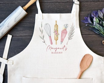 Puede incluir: Un delantal de color crema con un gráfico impreso de un batidor, un rodillo con detalles florales y una manga pastelera. El texto "Margaret's BAKERY" está impreso en rosa debajo de las ilustraciones. El delantal incluye un bolsillo frontal.