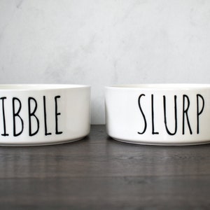 May include: Two white ceramic pet bowls with black lettering. The bowl on the left says "Nibble" and the bowl on the right says "Slurp".